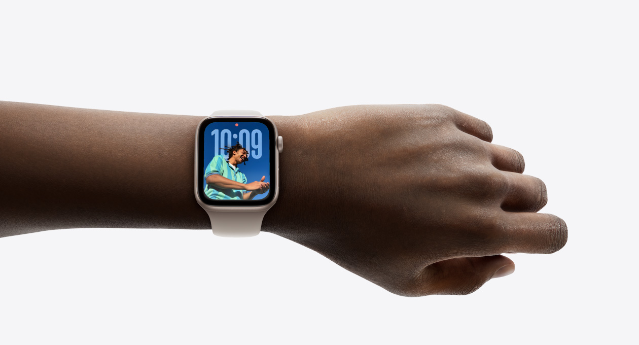ภาพระยะใกล้ของ Apple Watch สีเงินพร้อมสายที่ทำจากยางบนข้อมือของบุคคล, แสดงแอปเวลาในดีไซน์แบบ Liquid Glass ใหม่