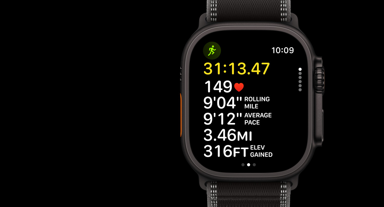 Apple Watch Ultra 3 med urkasse i sort titanium, appen Træning og Outdoor Run på skærmen, Handlingsknap i venstre side, Digital Crown og sideknap i højre side og Trail Loop-rem i sort/koksgrå