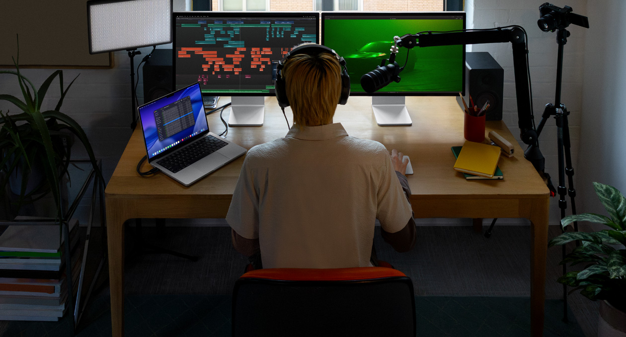 A person sitting at a desk with a mic and headphones in front of three devices, Mac Studio Display screen, audio editing tools, sound clips in different blocks of colour, a second Mac Studio Display screen showing, a moody shot of a car in the fog with green lighting, MacBook Pro 16 screen showing the Finder app on desktop showing Files for Final Cut Pro, dock, blue stylized desktop background