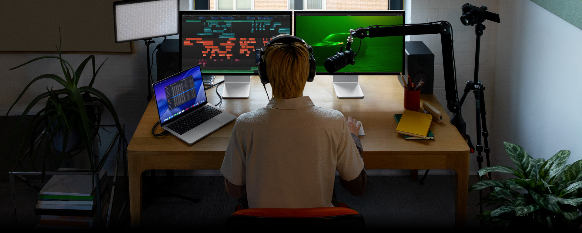 A person sitting at a desk with a mic and headphones in front of three devices, Mac Studio Display showing detailed audio editing tools, sound clips in different blocks of colour, a second Mac Studio Display screen showing a car with moody green lighting, a MacBook Pro 16 screen showing a long list of shots in a Final Cut Pro project