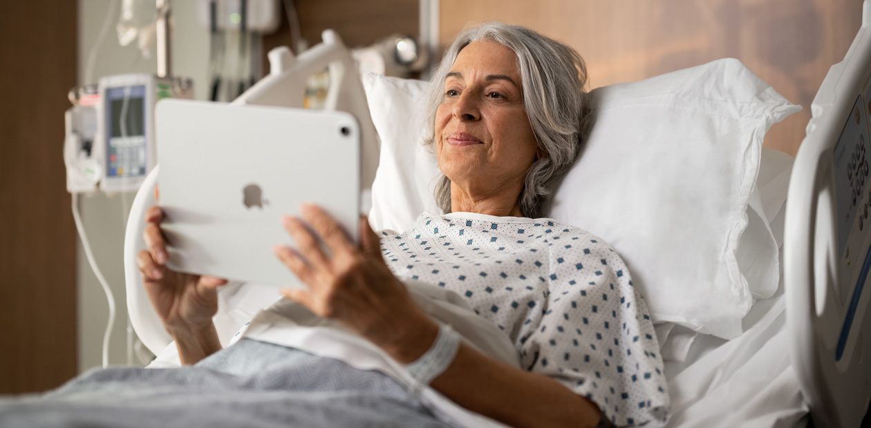 صورة لمريضة مستلقية على سرير المستشفى بينما تنظر إلى جهازها iPad‏.