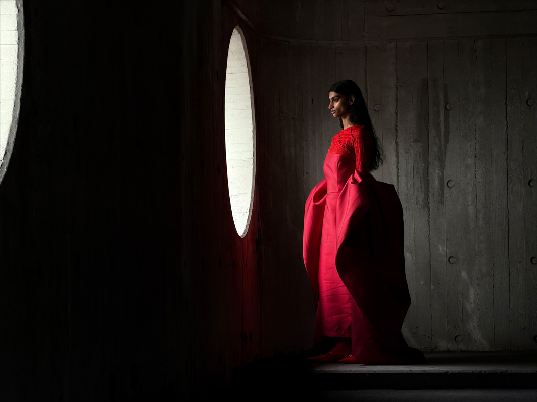 Eine Frau in einem langen roten Kleid steht in einem leeren Raum und wird fotografiert. Die Fotos App wechselt durch die Brennweiten, um die Möglichkeiten des Pro Kamera-Systems zu zeigen, hier: 100 mm